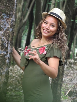 A person standing in a forested area, wearing a green dress with floral embroidery on the shoulders and a straw hat. The individual is smiling and holding glasses in one hand while leaning against a tree.