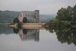 A stone chapel with a small tower is situated on a serene waterfront, reflecting in the still water. The building is adorned with a large American flag hanging from one side. Surrounding the chapel are trees and shrubs, while a hazy sky looms in the background, adding a sense of tranquility to the scene.