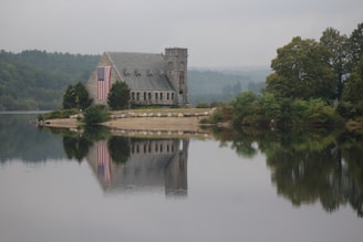 A stone chapel with a small tower is situated on a serene waterfront, reflecting in the still water. The building is adorned with a large American flag hanging from one side. Surrounding the chapel are trees and shrubs, while a hazy sky looms in the background, adding a sense of tranquility to the scene.