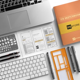 A neatly arranged workspace features a laptop, a UX sketchbook, a keyboard, drawing tools, stencil templates, and wireframe sketches for a mobile app design. The environment appears organized and intended for user experience design work.