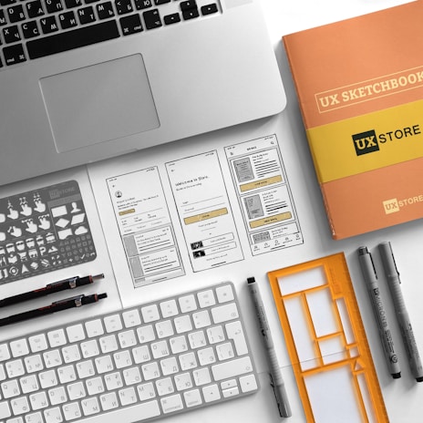 A neatly arranged workspace features a laptop, a UX sketchbook, a keyboard, drawing tools, stencil templates, and wireframe sketches for a mobile app design. The environment appears organized and intended for user experience design work.