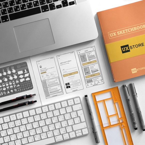 A neatly arranged workspace features a laptop, a UX sketchbook, a keyboard, drawing tools, stencil templates, and wireframe sketches for a mobile app design. The environment appears organized and intended for user experience design work.
