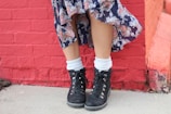 A person is standing in front of a red brick wall, wearing a long, flowing skirt with a paisley pattern and a pair of sturdy black hiking boots with white socks peeking out.