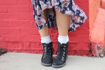 A person is standing in front of a red brick wall, wearing a long, flowing skirt with a paisley pattern and a pair of sturdy black hiking boots with white socks peeking out.