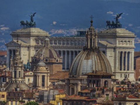 A cityscape featuring historical architecture with ornate domes and classical columns. The buildings are detailed, with a prominent large white structure adorned with statues of horses and chariots on its roof. The foreground includes smaller domed structures and traditional buildings with tiled roofs.