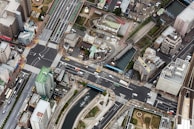 An aerial view of an urban landscape featuring a complex network of roads and intersections, surrounded by high-rise buildings and smaller structures. Vehicles can be seen navigating the streets, and a river runs parallel to one of the main roads. There are green spaces and pathways alongside the river.