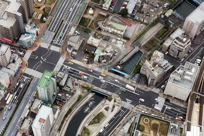 An aerial view of an urban landscape featuring a complex network of roads and intersections, surrounded by high-rise buildings and smaller structures. Vehicles can be seen navigating the streets, and a river runs parallel to one of the main roads. There are green spaces and pathways alongside the river.