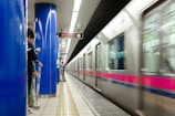 A train is moving quickly through an underground station, captured with a motion blur. People are standing on the platform, some with their phones out, next to bright blue support columns. An overhead sign displays train information in both Japanese and English.