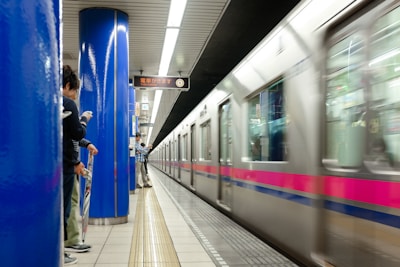 A train is moving quickly through an underground station, captured with a motion blur. People are standing on the platform, some with their phones out, next to bright blue support columns. An overhead sign displays train information in both Japanese and English.