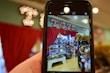 A phone held in the foreground captures a vibrant toy store display. The store features shelves filled with a variety of toys, including stuffed animals and boxes of games. Above the display, a bright marquee sign spells 'Toys' against a backdrop of red curtains.