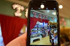 A phone held in the foreground captures a vibrant toy store display. The store features shelves filled with a variety of toys, including stuffed animals and boxes of games. Above the display, a bright marquee sign spells 'Toys' against a backdrop of red curtains.
