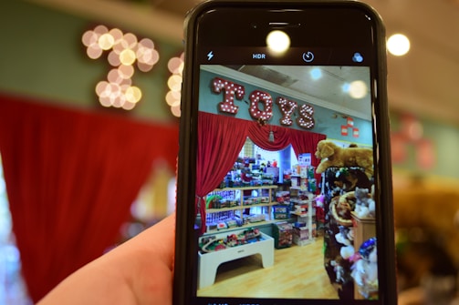 A phone held in the foreground captures a vibrant toy store display. The store features shelves filled with a variety of toys, including stuffed animals and boxes of games. Above the display, a bright marquee sign spells 'Toys' against a backdrop of red curtains.