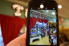 A phone held in the foreground captures a vibrant toy store display. The store features shelves filled with a variety of toys, including stuffed animals and boxes of games. Above the display, a bright marquee sign spells 'Toys' against a backdrop of red curtains.