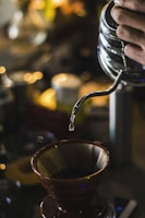 A softly lit scene of a hand pouring coffee from a dripper into a rounded ceramic mug.