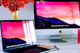 A workspace featuring an Apple iMac and a MacBook Pro, both displaying the same screen wallpaper of a mountainous landscape at sunset. A smartphone and a white keyboard are placed on a white desk. A vase with vibrant orange flowers is visible on the left.