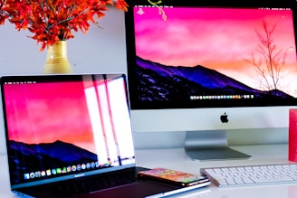 A workspace featuring an Apple iMac and a MacBook Pro, both displaying the same screen wallpaper of a mountainous landscape at sunset. A smartphone and a white keyboard are placed on a white desk. A vase with vibrant orange flowers is visible on the left.