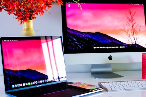 A workspace featuring an Apple iMac and a MacBook Pro, both displaying the same screen wallpaper of a mountainous landscape at sunset. A smartphone and a white keyboard are placed on a white desk. A vase with vibrant orange flowers is visible on the left.