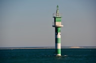 A green and white striped navigation buoy is situated in calm, open water with a distant sandy shoreline. The structure is topped with a small platform and a conical green finial.