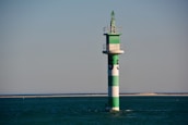 A green and white striped navigation buoy is situated in calm, open water with a distant sandy shoreline. The structure is topped with a small platform and a conical green finial.