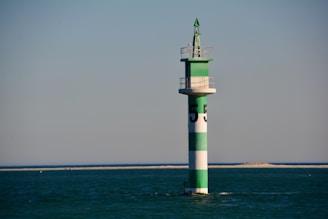 A green and white striped navigation buoy is situated in calm, open water with a distant sandy shoreline. The structure is topped with a small platform and a conical green finial.