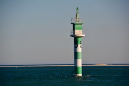 A green and white striped navigation buoy is situated in calm, open water with a distant sandy shoreline. The structure is topped with a small platform and a conical green finial.