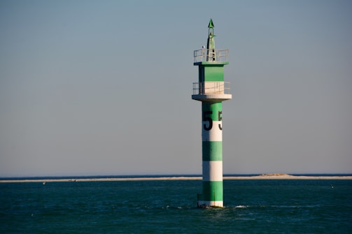 A green and white striped navigation buoy is situated in calm, open water with a distant sandy shoreline. The structure is topped with a small platform and a conical green finial.