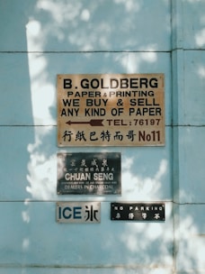 A series of signs are displayed on a light blue wall, featuring English and Chinese text. The top sign advertises paper and printing services, the middle sign mentions a charcoal dealer, and the lower signs include an 'ICE' label and a 'NO PARKING' notice.