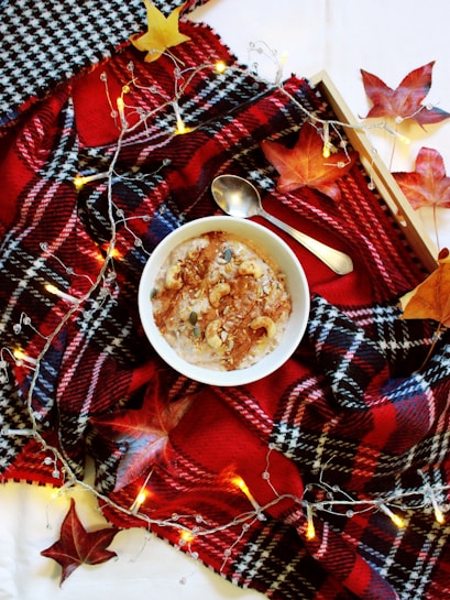 A cozy scene featuring a bowl of oatmeal topped with nuts and seeds, resting on a red and black plaid blanket. Decorative autumn leaves and a string of warm fairy lights add an autumnal touch. A spoon lies next to the bowl, enhancing the inviting atmosphere.