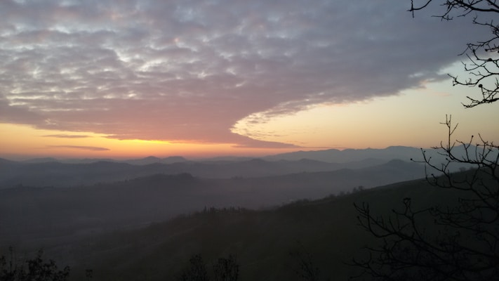 A serene landscape showcases a gentle sunrise or sunset with the sky painted in soft hues of orange and blue. Rolling hills and distant mountains create a layered effect in the background, with a misty ambience enveloping the scene. Bare tree branches slightly frame the right side of the image, adding depth and framing to the composition.