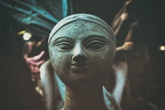 A close-up view of a clay sculpture depicting a serene human face with closed eyes and a slight smile. The background is dimly lit, with other partially visible sculptures, creating an atmosphere of mystique and focus on the central figure.