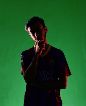 A person stands in silhouette against a vivid green background, wearing a dark sports jersey with colored stripes and a visible emblem. The lighting highlights the edges of the figure, casting shadows across their face.