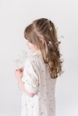 A young girl with long, curly hair adorned with small white flowers is wearing a light-colored dress with small floral patterns. She is holding delicate white flowers in her hands.