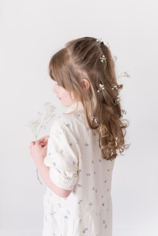 A young girl with long, curly hair adorned with small white flowers is wearing a light-colored dress with small floral patterns. She is holding delicate white flowers in her hands.