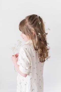 A young girl with long, curly hair adorned with small white flowers is wearing a light-colored dress with small floral patterns. She is holding delicate white flowers in her hands.