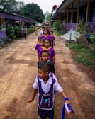 A group of children in school uniforms is lined up in an orderly manner along a narrow path between two buildings. The children are dressed in purple and white uniforms and appear to be in a school setting. There are adult figures and trees in the background.