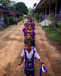 A group of children in school uniforms is lined up in an orderly manner along a narrow path between two buildings. The children are dressed in purple and white uniforms and appear to be in a school setting. There are adult figures and trees in the background.