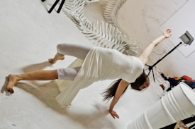 A person is performing a dynamic dance pose on a white floor, wearing a white outfit. Surrounding them are stacked white chairs creating an artistic backdrop. There is a spotlight on a stand and a white barrel in the foreground.