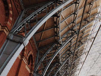 Architectural elements of a large indoor structure with intricate metal beams and red brick walls. The ceiling features a series of arched trusses, displaying a mix of industrial and historic design.