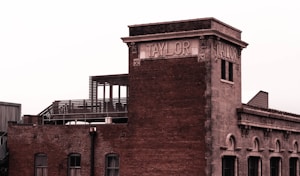 A historic brick building with a sign reading 'TAYLOR' on the upper section. The building has arched windows and a rooftop terrace with metal railings. The architecture appears to be from an older era, conveying a sense of nostalgia.