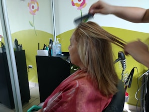A person is seated in a hair salon with a cape around their shoulders. Another individual is styling their hair using a comb. The salon has bright decorations on the wall, including flowers, and there are various hair styling tools and products on a counter.