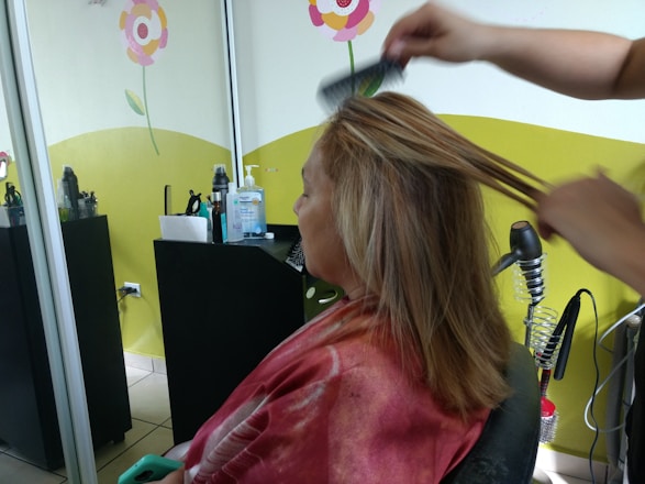 A person is seated in a hair salon with a cape around their shoulders. Another individual is styling their hair using a comb. The salon has bright decorations on the wall, including flowers, and there are various hair styling tools and products on a counter.