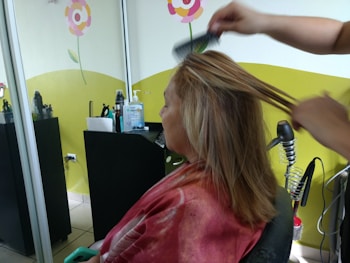 A person is seated in a hair salon with a cape around their shoulders. Another individual is styling their hair using a comb. The salon has bright decorations on the wall, including flowers, and there are various hair styling tools and products on a counter.