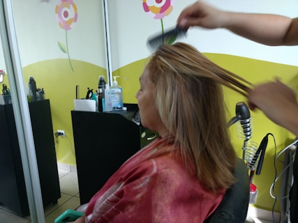 A person is seated in a hair salon with a cape around their shoulders. Another individual is styling their hair using a comb. The salon has bright decorations on the wall, including flowers, and there are various hair styling tools and products on a counter.