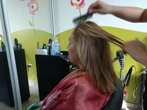 A person is seated in a hair salon with a cape around their shoulders. Another individual is styling their hair using a comb. The salon has bright decorations on the wall, including flowers, and there are various hair styling tools and products on a counter.