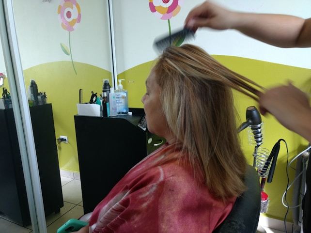 A person is seated in a hair salon with a cape around their shoulders. Another individual is styling their hair using a comb. The salon has bright decorations on the wall, including flowers, and there are various hair styling tools and products on a counter.