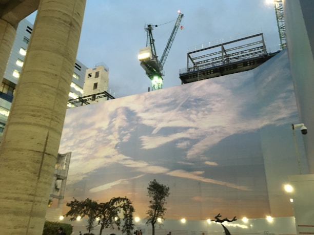 A large fabric printed with a serene sky and clouds covers the side of a construction site. The scene includes cranes above the printed image, with a mix of high-rise buildings and exposed frameworks visible in the background. Small trees and shrubbery are present at the base, illuminated by several lights.