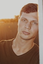 A young man with short brown hair and clear skin is leaning against a surface. The play of sunlight creates a warm glow on his face, and he appears relaxed. His gaze is directed towards the camera, surrounded by a softly blurred background that suggests an outdoor setting.