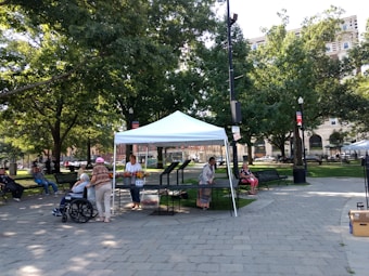 A small outdoor market stall is set up under a white canopy in a park. A person is arranging flowers priced at $10 on a table. Several people are sitting on nearby benches, including one in a wheelchair. The area is surrounded by tall trees providing shade and there are large buildings in the background.