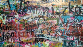 A vibrant and colorful wall covered in numerous layers of graffiti and street art, featuring various words, phrases, and symbols in a chaotic and expressive display. The artwork blends a multitude of colors and styles, creating an intricate tapestry of urban expression.