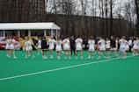 A group of female athletes in white sports uniforms are positioned on a green sports field. The players are wearing white shorts and jerseys with numbers on their backs, accompanied by sports shoes. They appear to be gathering in a somewhat circular formation, possibly preparing for a game or practice. Behind them, there is a white tent with several people inside, and a forested area with leafless trees, suggesting it is a cool season.
